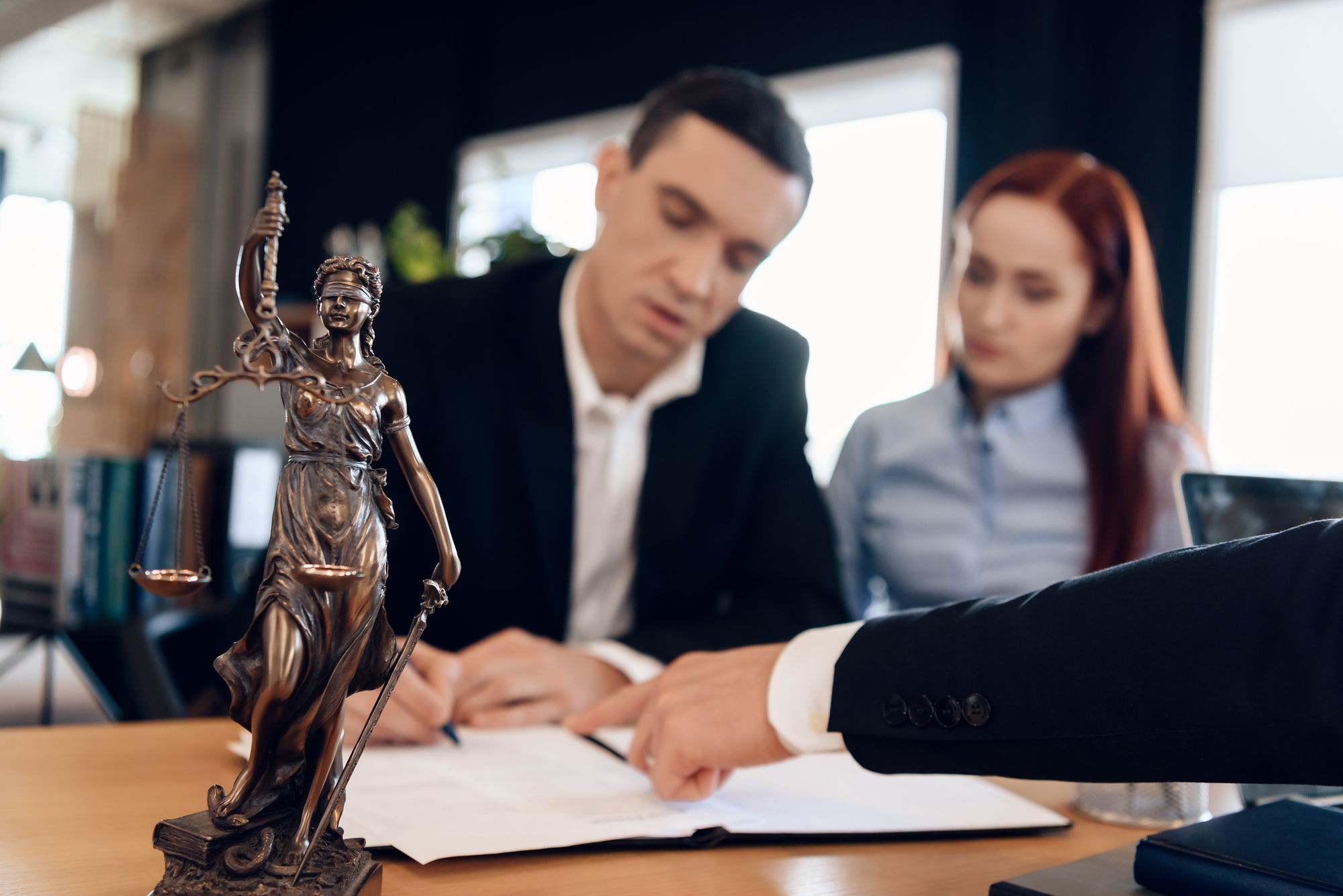 Statue of Themis holds scales of justice. In unfocused background, adult man signs documents.